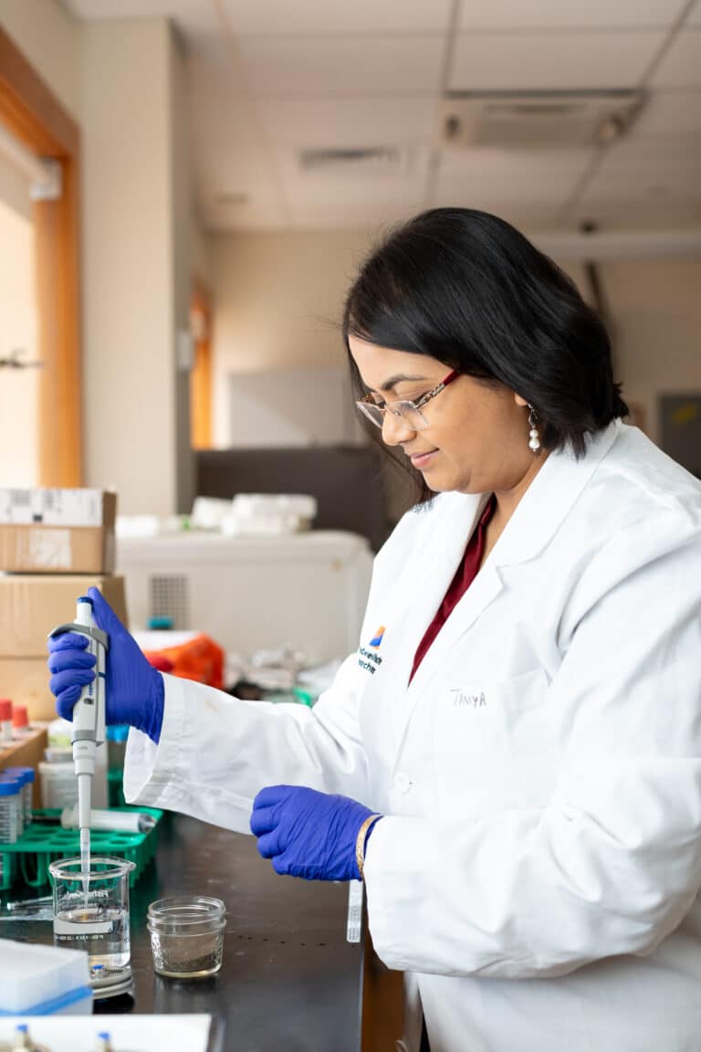 Woman with dark hair and glasses, wearing a white lab coat and blue gloves, distributing samples at a laboratory bench.