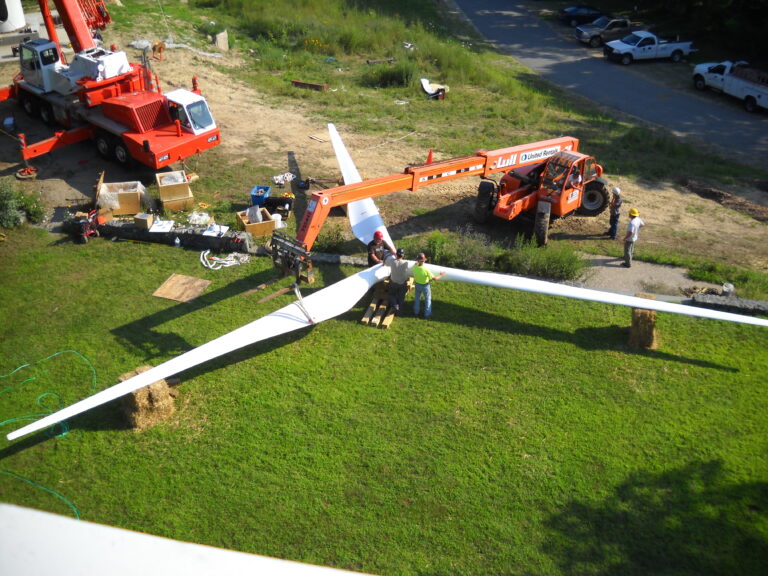 Workers stand around the large blades of a wind turbine laying on the lawn, preparing for installation.