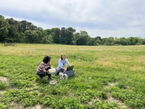 Two young women sit in a field, adjusting scientific equipment attached to a laptop.