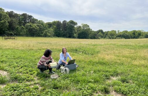 Two young women sit in a field, adjusting scientific equipment attached to a laptop.