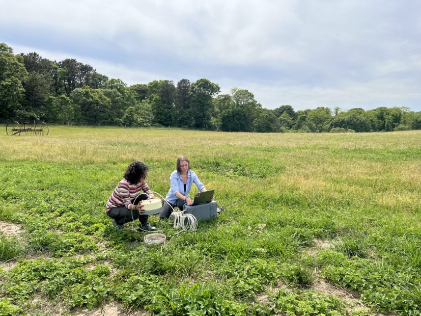 Two young women sit in a field, adjusting scientific equipment attached to a laptop.