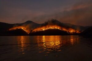 The glowing edge of a fire crawls across the topography of a hill, reflection of the glow shown in the water in the foreground