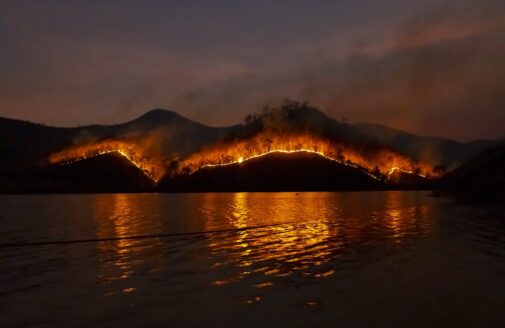 The glowing edge of a fire crawls across the topography of a hill, reflection of the glow shown in the water in the foreground