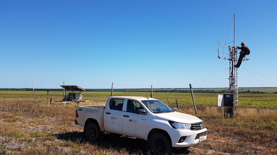 a truck stands in the foreground while a person works at the top of a tower 3-4 times their height to adjust scientific instruments