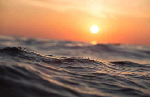 The orange sun sits low in the sky over an ocean wave in the foreground