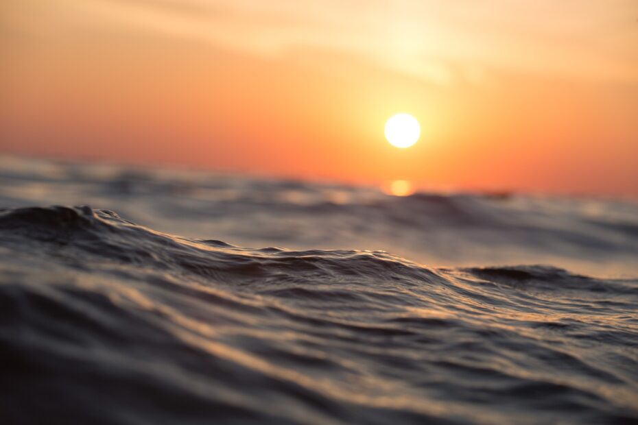 The orange sun sits low in the sky over an ocean wave in the foreground