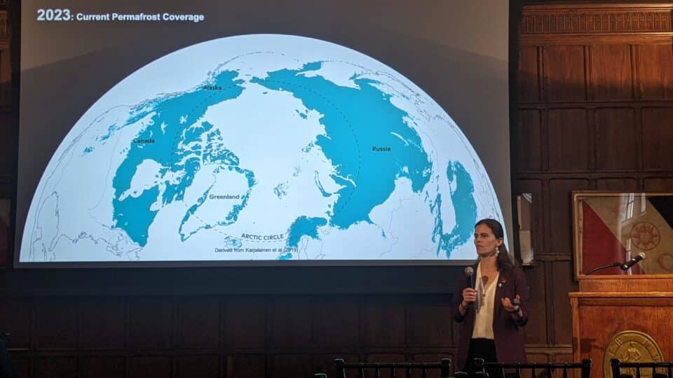 a woman speaks into a microphone in front of a large projected map of the northern hemisphere, showing permafrost extent