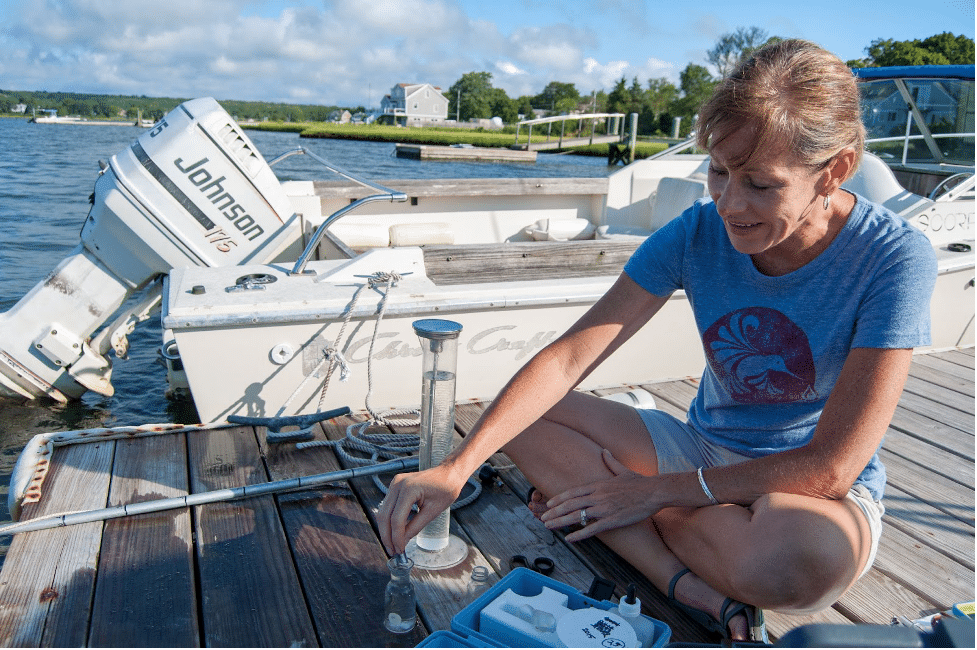 a woman sits on a boardwalk next to a tube of water, using a dropper to take water out of another small container