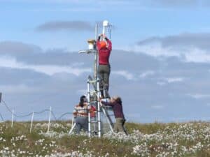 two researchers hold a ladder steady for a third researcher who is working with equipment at the top of a mid-size tower in Alaska