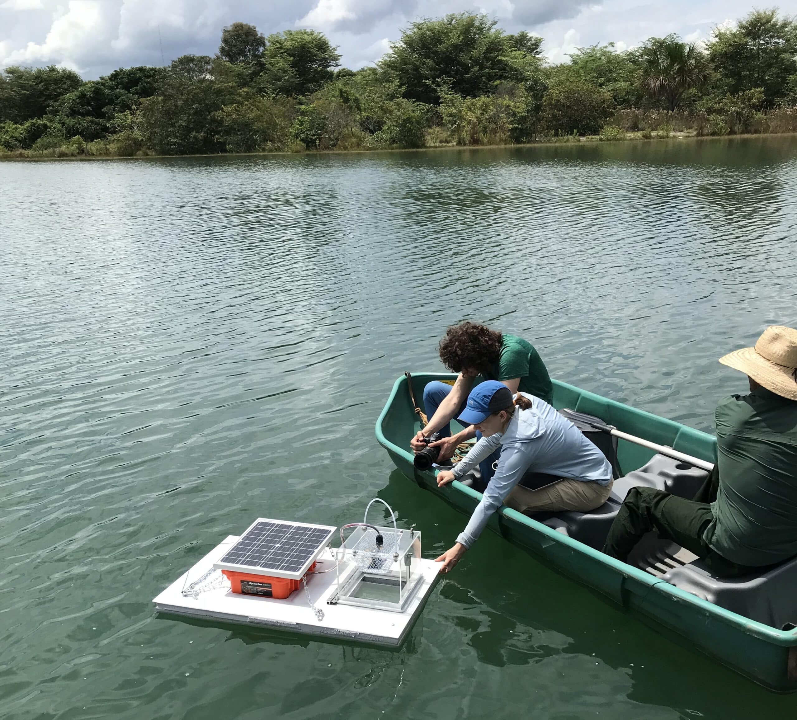 a researcher sits in a boat with two other people, and leans over the side to adjust a scientific instrument floating on a white piece of flat material on top of the water