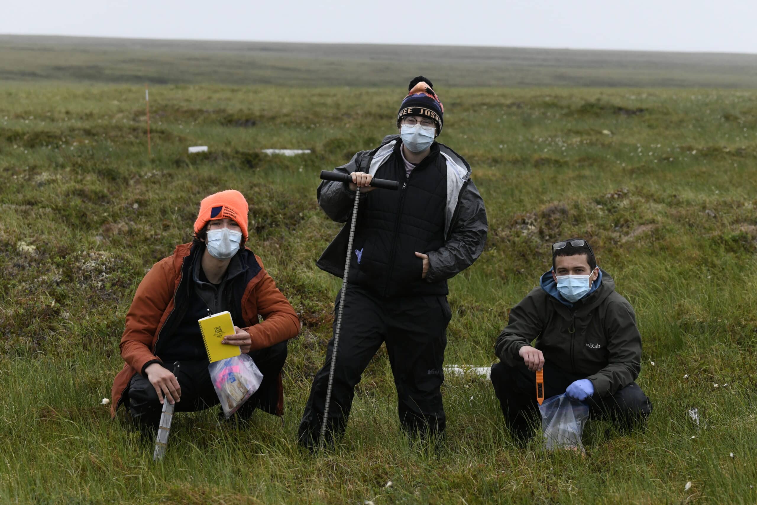 three young researchers wearing face masks stand in the arctic tundra, surrounded by green grass. Each holds a different piece of science equipment, including a field journal, a manual auger, and a thermometer.