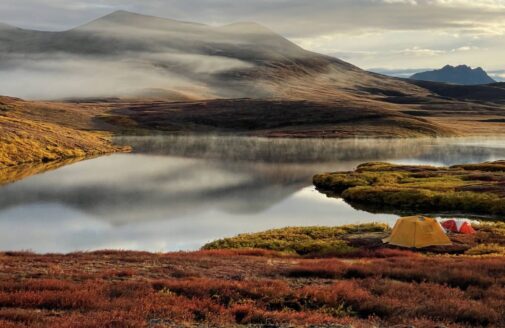 richly colored arctic landscape with orange grasses, a few tents in the foreground, and a light blue lake reflecting clouds of a dynamic array of greys