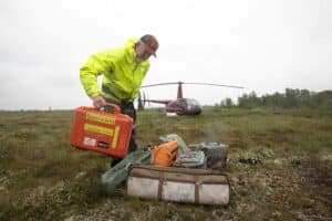 Dr. R. Max Holmes adds an orange tote labeled "Science on the Fly" to a pile of equipment, with a helicopter in the background