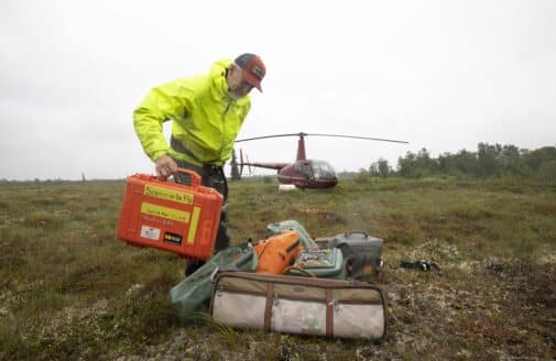 Dr. R. Max Holmes adds an orange tote labeled "Science on the Fly" to a pile of equipment, with a helicopter in the background