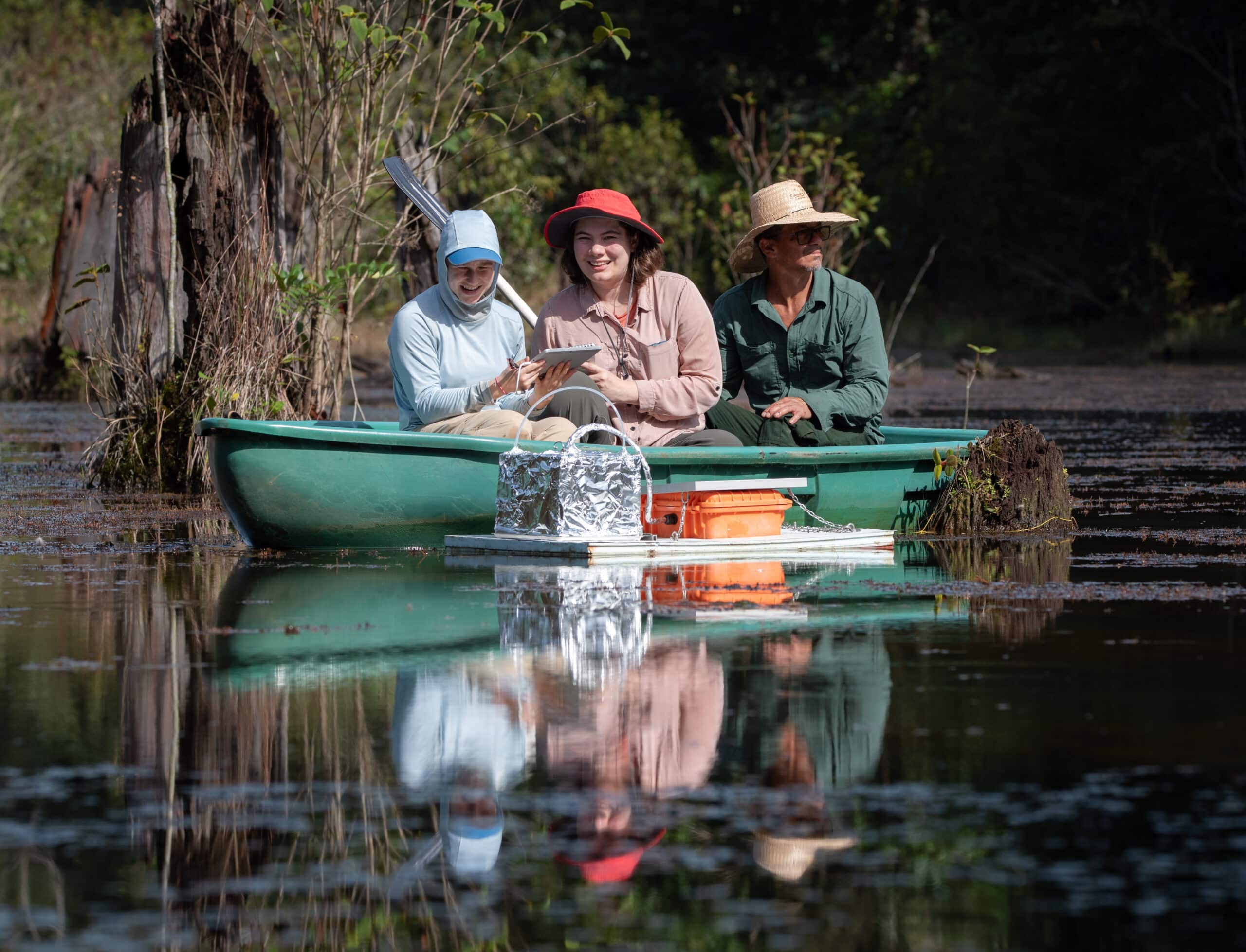 three people sit on a boat looking at a floating methane chamber