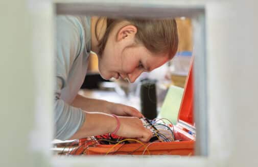 picture through a cut out in an aluminum sheet, Zoe dietrich works on wires in an orange box