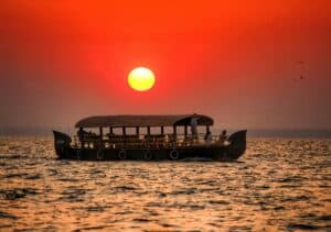 a long boat with a roof over it floats on the water below a red sun in a hazy sky