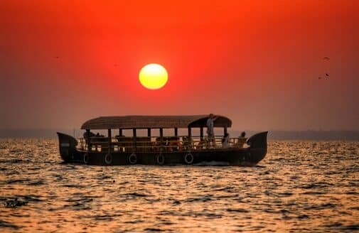 a long boat with a roof over it floats on the water below a red sun in a hazy sky