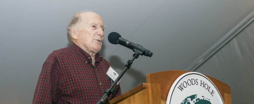 George Woodwell stands at a podium speaking at an event under a tent