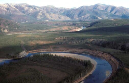 the bending arm of a river cuts through green grassy land with evergreen trees, mountains looming in the background