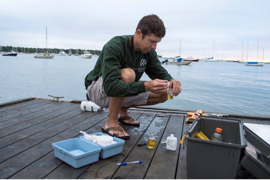 a man crouches on a dock by the water, using a dropper to get a small amount of yellow liquid out of a bottle.