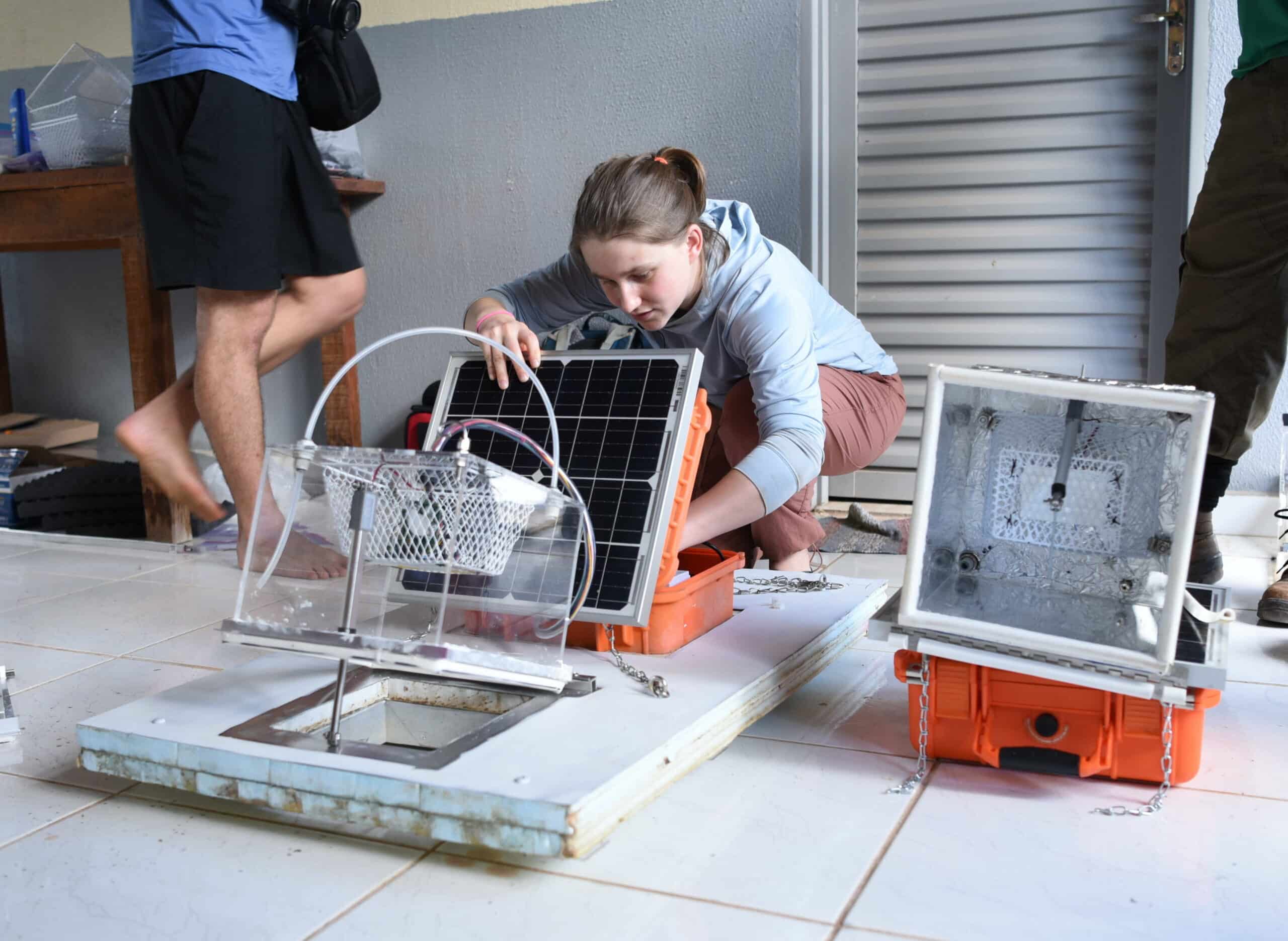 Zoe Dietrich crouches near her autonomous methane chamber, a clear box hooked up to wires on a white flat platform. She is working in an open orange box with a solar panel on top