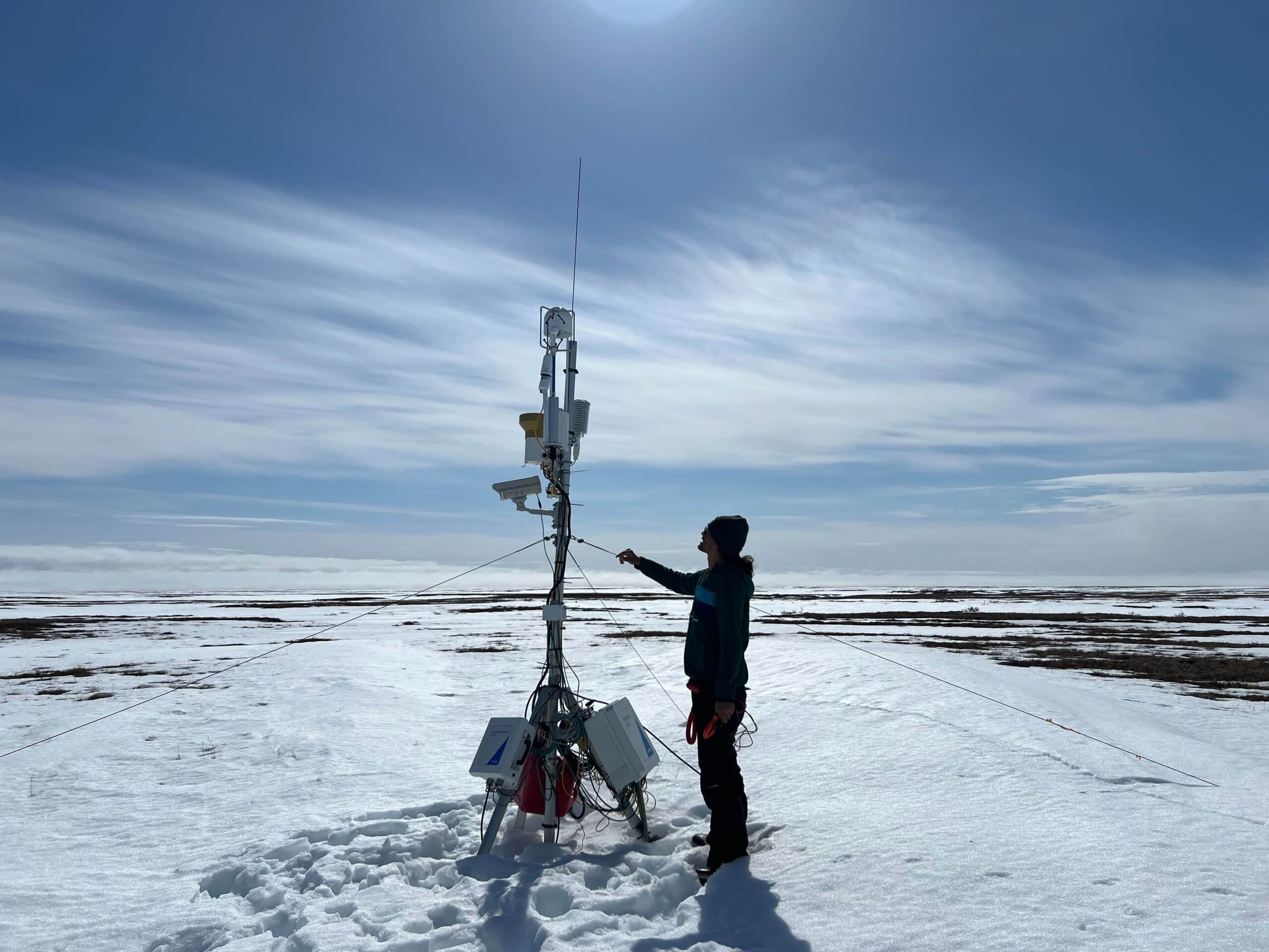 a researcher stands on a snowy expanse next to a slim tower with scientific instruments on it