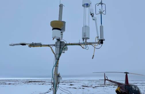 scientific instruments mounted to a slim pole in the foreground, with a helicopter on a snowy field in the background