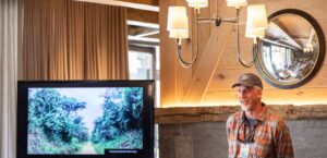 Dr. Max Holmes dressed in a plaid shirt and baseball cap, speaking next to a screen with an image of a forest displayed