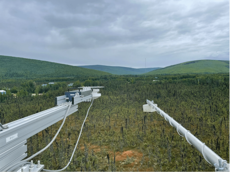 a view of a sparse boreal forest from a tower. Two white beams stretch into the photo with scientific instruments mounted on the ends