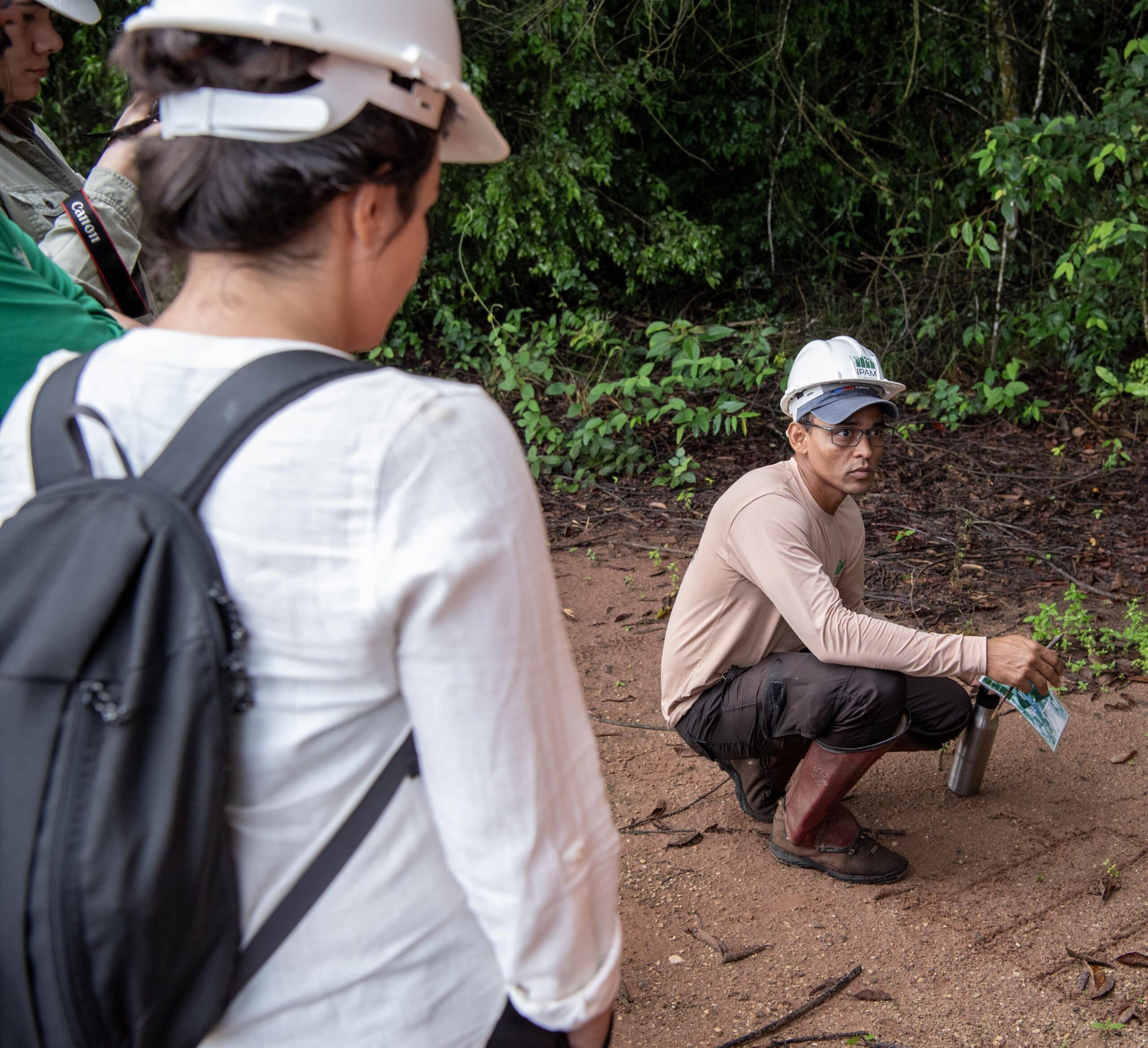 a researcher crouches on the ground, explaining something to two other people. all are wearing white hard hats