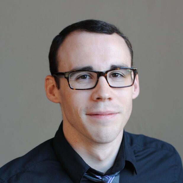 photo portrait of Joey Eschrich, a white man wearing glasses, a black shirt, and a blue tie