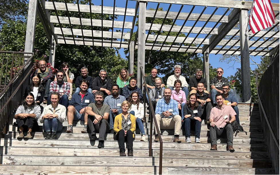 a group photo of people sitting on wooden stairs outdoors, underneath a pergola