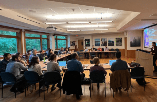 a group of researchers sits at a horseshoe-shaped table, looking at a projector screen