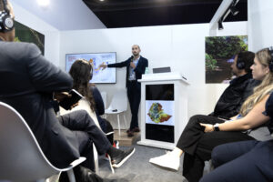 a man presents to a group of seated people, pointing to a map on a screen. The people listening are wearing headphones.