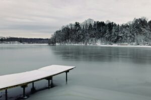 a snow-covered dock juts out into a frozen lake, with evergreen trees on the far shore