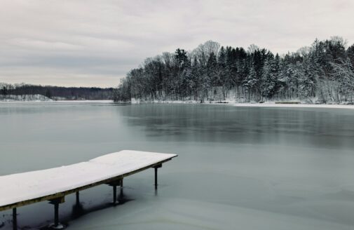 a snow-covered dock juts out into a frozen lake, with evergreen trees on the far shore